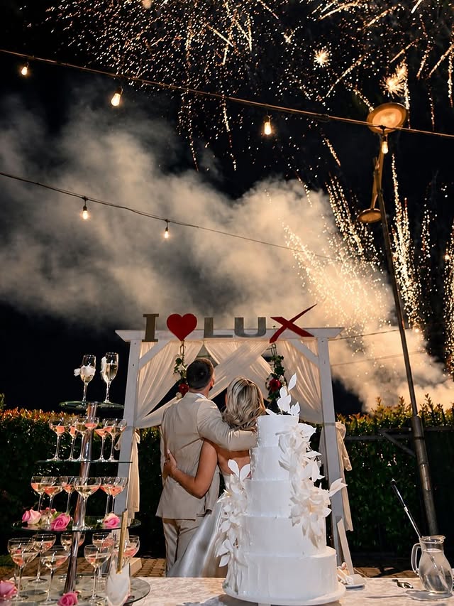 Mladenci zagrljeni leđima okrenuti prema vatrometu iznad I ❤️ LUX konstrukcije, sa svadbeneom tortom i čašama šampanjca u prvom planu na venčanju u New Lux Events Center-u